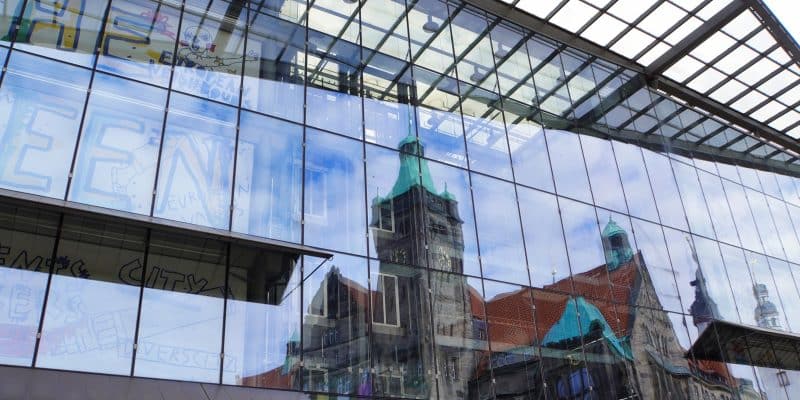 In der modernen Glasfassade eines Gebäudes in Chemnitz spiegelt sich das historische Rathaus mit seinem markanten grünen Turm und den roten Ziegeldächern unter einem leicht bewölkten Himmel.