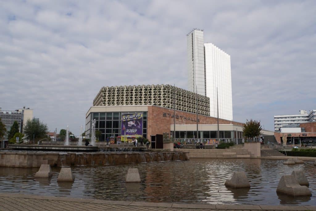 Die Stadthalle Chemnitz mit ihrer markanten Wabenfassade steht hinter einem großen Brunnenbecken mit kleinen Wasserfällen, während im Hintergrund das hohe, weiße Gebäude des Dorint Kongresshotels aufragt.