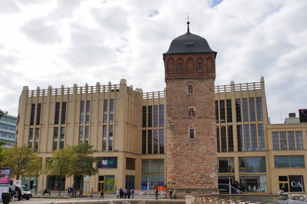 Ein historischer, rötlicher Backsteinturm, der Rote Turm in Chemnitz, steht vor einem modernen, mehrstöckigen Einkaufszentrum mit großen Glasfronten und heller Fassade. Vor den Gebäuden beleben Passanten einen gepflasterten Platz mit Bäumen und einem Brunnen im Vordergrund.