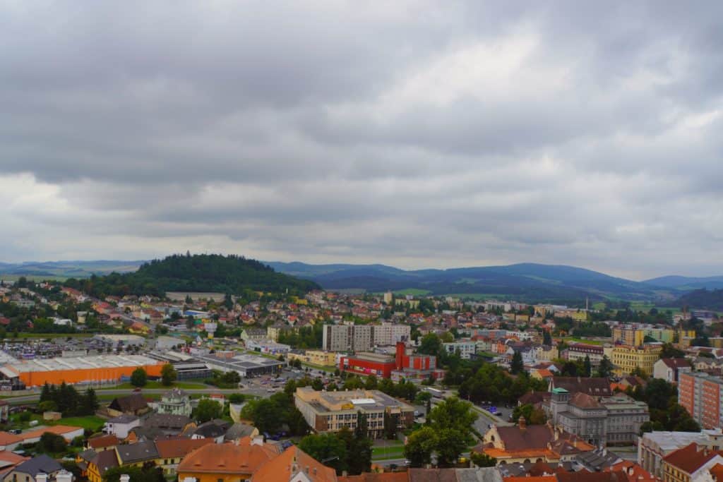 Blick vom Turm über die Stadt Klattau mit ihren bunten Häusern, Wohnblöcken und Gewerbegebieten vor einer bewaldeten Hügellandschaft unter bewölktem Himmel.