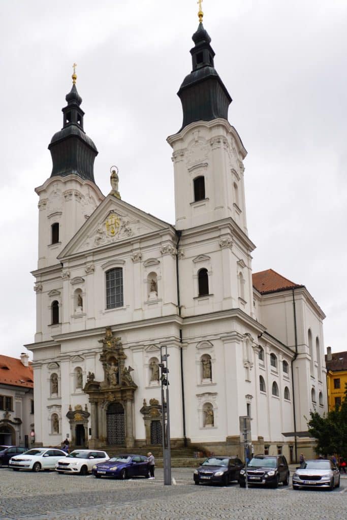 Die prachtvolle weiße Fassade der Jesuitenkirche in Klattau mit ihren zwei markanten Zwiebeltürmen und barocken Skulpturen ragt hinter einer Reihe parkender Autos an einem gepflasterten Platz empor.