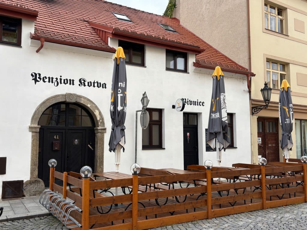 Die Außenansicht der Pension Kotva in Klattau zeigt ein weißes Gebäude mit rotem Ziegeldach, einer hölzernen Außenterrasse mit Sitzbänken und Sonnenschirmen sowie einem markanten Rundbogenportal.