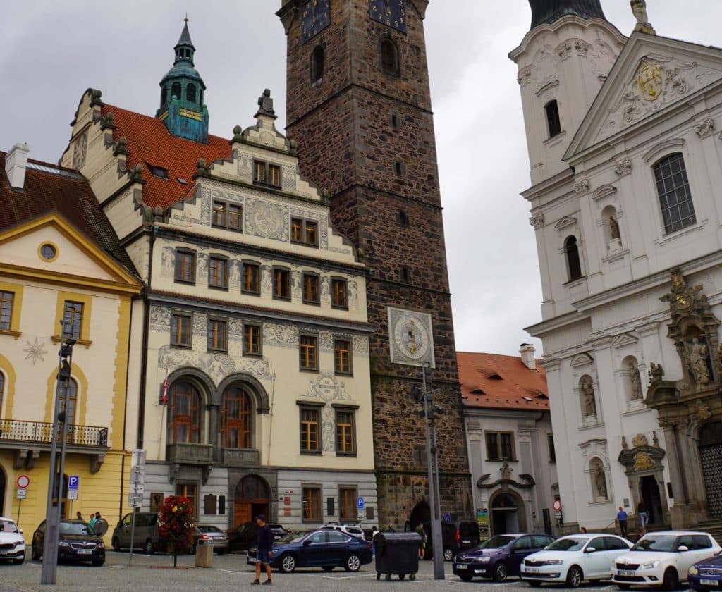 Blick auf das historische Rathaus von Klattau mit seiner kunstvoll verzierten Fassade, flankiert vom massiven Schwarzen Turm aus dunklem Stein und einer weißen Barockkirche an einem belebten Platz mit parkenden Autos.