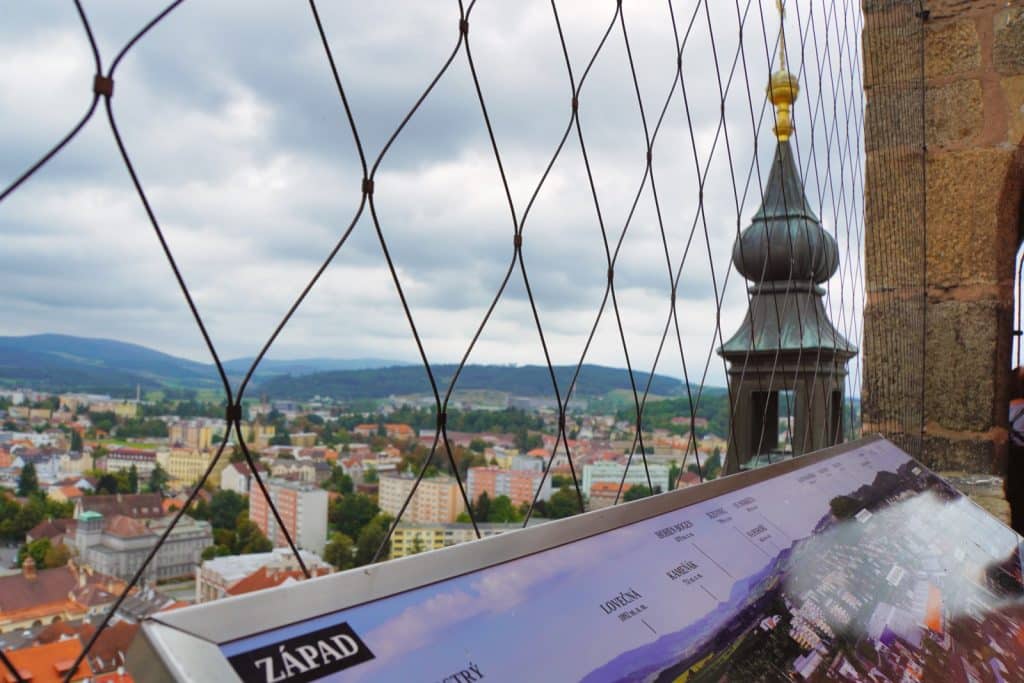 Blick vom Schwarzen Turm in Klattau durch ein Sicherheitsnetz auf das Stadtpanorama und eine Informationstafel mit der Aufschrift „ZÁPAD“ (Westen), während im Vordergrund eine Turmspitze mit goldener Kugel aufragt.