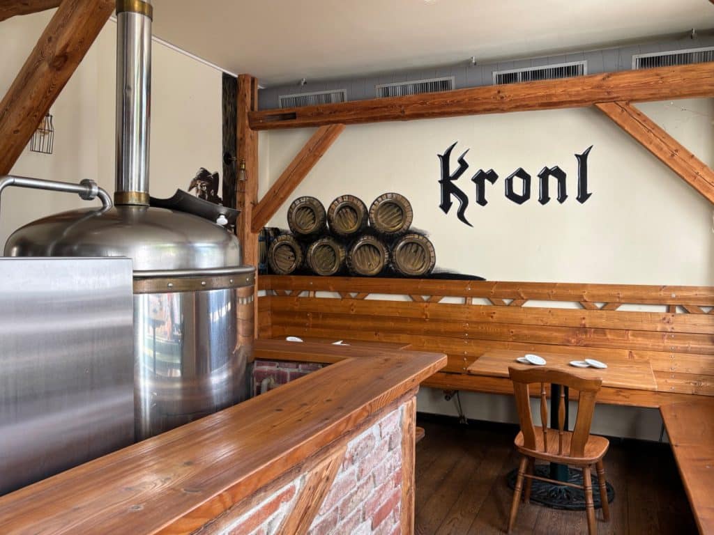 Innenansicht der Brauerei mit einem glänzenden Sudkessel aus Edelstahl im Vordergrund und einem rustikalen Sitzbereich aus Holz unter dem Schriftzug „Kronl“ an der Wand.
