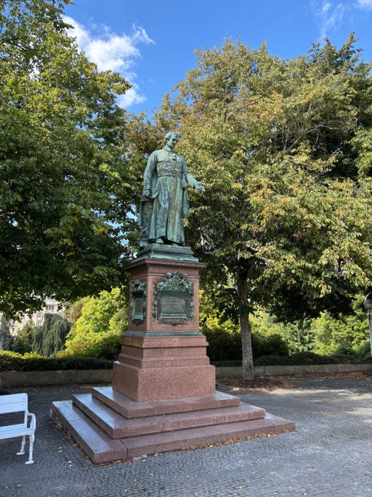 Sehenswürdigkeiten in Marienbad: Das Denkmal für Abt Kaspar Karl Reitenberger zeigt eine bronzene Statue des Klostergründers in langem Gewand auf einem hohen, rötlichen Granitsockel, umgeben von grünen Bäumen.