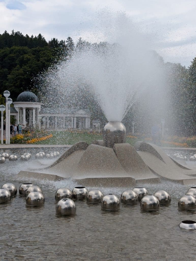 Die Singende Fontäne eine der Sehenswürdigkeiten in Marienbad sprüht eine hohe Wasserfontäne aus einer zentralen Steinskulptur, umgeben von zahlreichen im Wasser treibenden Metallkugeln und einer historischen Kolonnade im Hintergrund.