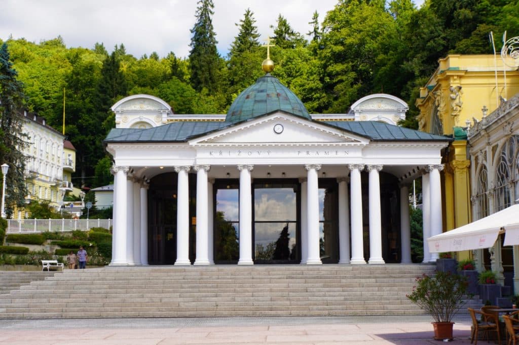 Das Bild zeigt die klassizistische Kolonnade der Kreuzquelle (Křížový pramen) in Marienbad mit ihren weißen Säulen, einer markanten grünen Kuppel mit goldenem Kreuz und einer breiten Freitreppe im Vordergrund. Im Hintergrund erstreckt sich eine bewaldete Hügellandschaft hinter dem historischen Kurgebäude.