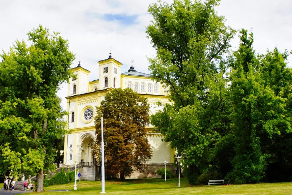 Die gelb-weiße römisch-katholische Mariä-Himmelfahrt-Kirche in Marienbad mit ihren zwei Türmen und einer großen Fensterrosette, umgeben von hohen grünen Bäumen und einer gepflegten Parkanlage.