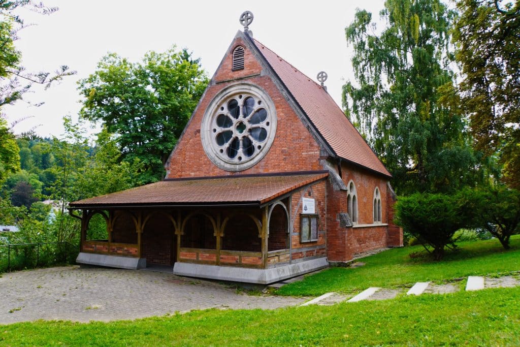Sehenswürdigkeiten in Marienbad: Eine kleine, backsteinerne Kirche mit einem markanten runden Rosettenfenster an der Giebelseite und einem hölzernen Vorbau steht auf einer grünen Wiese.
