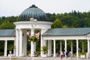 Die weiße Kolonnade in Marienbad mit ihrer markanten dunklen Kuppel und den korinthischen Säulen erstreckt sich vor einer bewaldeten Hügelkette. Im Vordergrund beleben Spaziergänger den gepflasterten Platz, der mit Blumenampeln und Palmen in Kübeln geschmückt ist.