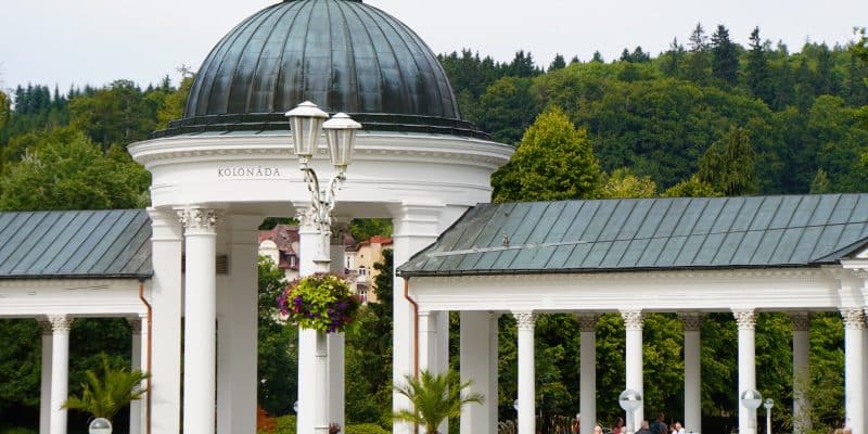 Die weiße Kolonnade in Marienbad mit ihrer markanten dunklen Kuppel und den korinthischen Säulen erstreckt sich vor einer bewaldeten Hügelkette. Im Vordergrund beleben Spaziergänger den gepflasterten Platz, der mit Blumenampeln und Palmen in Kübeln geschmückt ist.