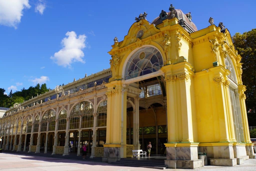 Sehenswürdigkeiten in Marienbad: Die prachtvolle, gelbe gusseiserne Kolonnade in Marienbad erstreckt sich mit ihren filigranen Rundbögen und kunstvollen Verzierungen unter einem strahlend blauen Himmel.