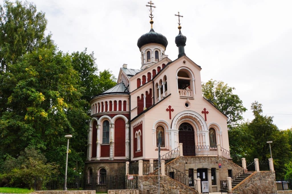 Sehenswürdigkeiten in Marienbad: Die russisch-orthodoxe Kirche des Heiligen Wladimir präsentiert sich als prachtvoller Bau mit rosa und roten Fassadenelementen, charakteristischen Zwiebeltürmen und einer steinernen Außentreppe vor bewaldetem Hintergrund.