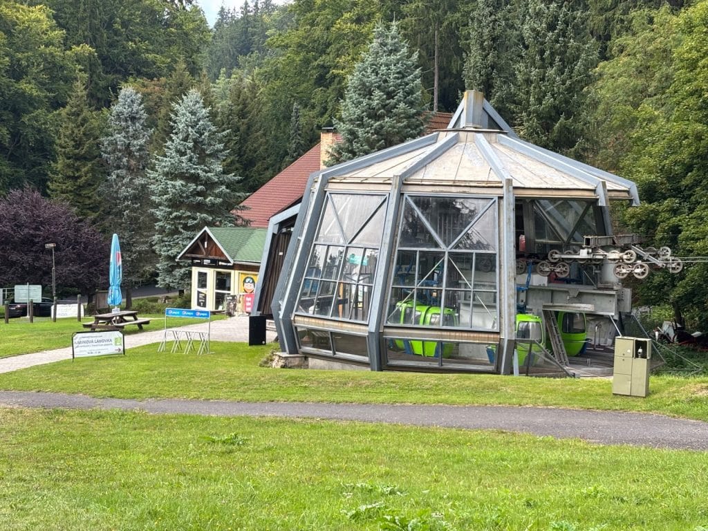 Die Talstation der Seilbahn in Marienbad mit ihren markanten, verglasten Kabinen in leuchtendem Grün vor einer bewaldeten Hügelkulisse.