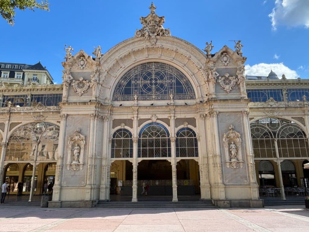 Das reich verzierte Portal der gusseisernen Kolonnade in Marienbad zeigt eine prachtvolle neobarocke Architektur mit filigranen Glasfronten, Skulpturen und einem großen Rundbogen unter blauem Himmel.