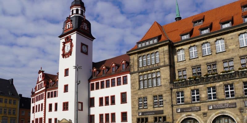 Das Alte Rathaus in Chemnitz, Sachsen, mit seinem markanten weißen Uhrturm und roten Verzierungen neben einem historischen Sandsteingebäude am Marktplatz unter blauem Himmel.