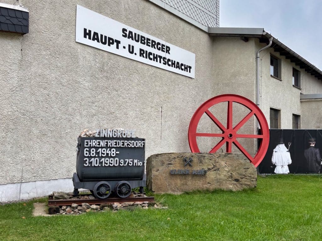 Außenansicht des Besucherbergwerks Zinngrube Ehrenfriedersdorf in Sachsen mit einem historischen Förderwagen auf Schienen, einem großen roten Förderrad und einer Gedenktafel am Gebäude.