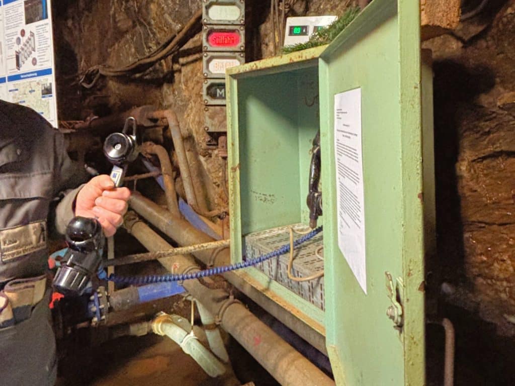 Ein Bergmann hält den Hörer eines historischen Grubentelefons in einem Stollen der Zinngrube Ehrenfriedersdorf in Sachsen, das in einem hellgrünen Metallkasten an der Felswand montiert ist.