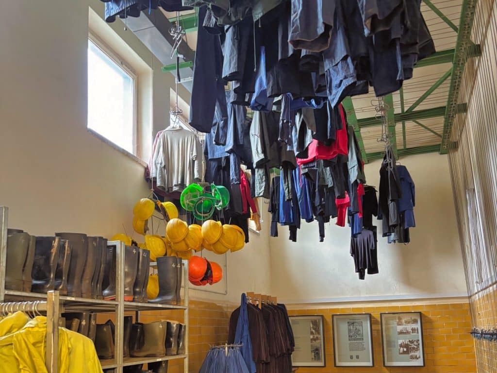 In einer historischen Kaue in Ehrenfriedersdorf, Sachsen, hängen zahlreiche Arbeitsanzüge an Ketten unter der hohen Decke, während darunter gelbe Schutzhelme und Regale mit Gummistiefeln angeordnet sind.