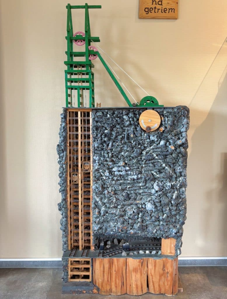 Ein detailliertes Modell eines Bergbau-Schachtes aus Ehrenfriedersdorf in Sachsen zeigt einen grünen Förderturm über einem Querschnitt durch das Gestein mit hölzernen Förderkörben und Schienenanlagen unter Tage.