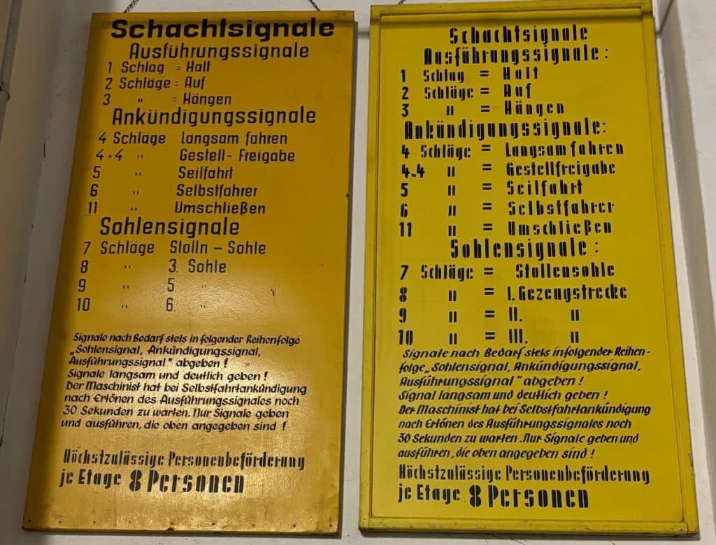 Zwei gelbe Hinweistafeln aus Ehrenfriedersdorf in Sachsen zeigen unter der Überschrift „Schachtsignale“ eine detaillierte Liste von Klopf- oder Glockensignalen für den Bergbau, unterteilt in Ausführungs-, Ankündigungs- und Sohlensignale. Die Tafeln enthalten zudem Sicherheitsanweisungen für den Maschinisten und geben die maximale Personenbeförderung pro Etage mit 8 Personen an.