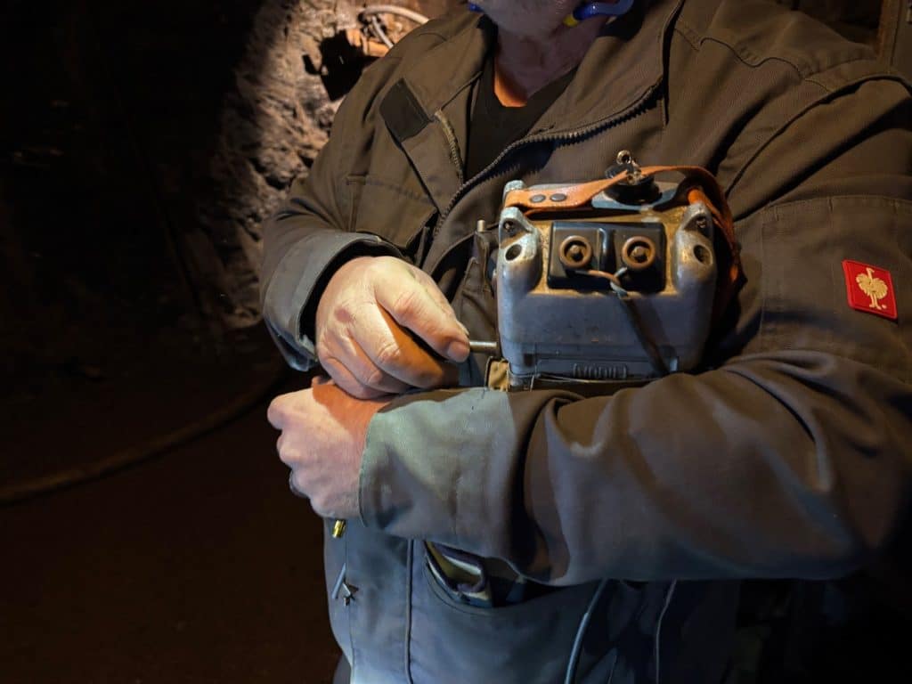 Ein Bergmann in Arbeitskleidung hält in einem Stollen in Ehrenfriedersdorf, Sachsen, eine historische, metallene Sprengvorrichtung vor seiner Brust und bedient einen seitlichen Hebel.