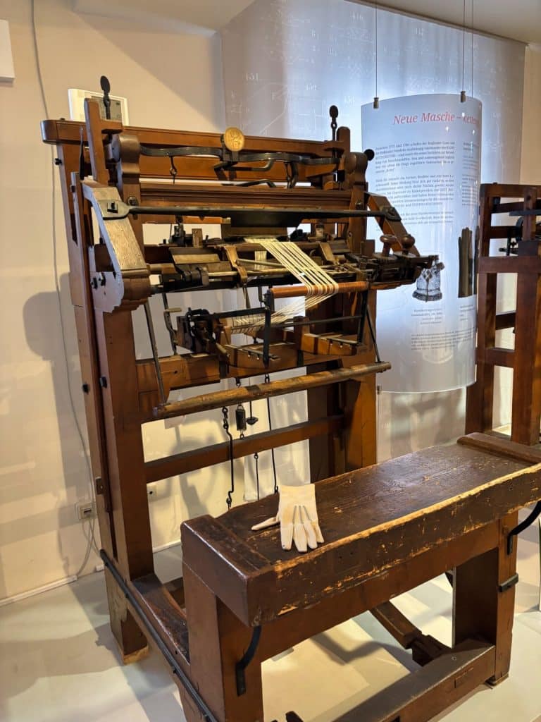 Ein historischer hölzerner Wirkstuhl zur Handschuhherstellung im Esche-Museum in Limbach-Oberfrohna, Sachsen, mit einem weißen Handschuh auf der Sitzbank im Vordergrund.