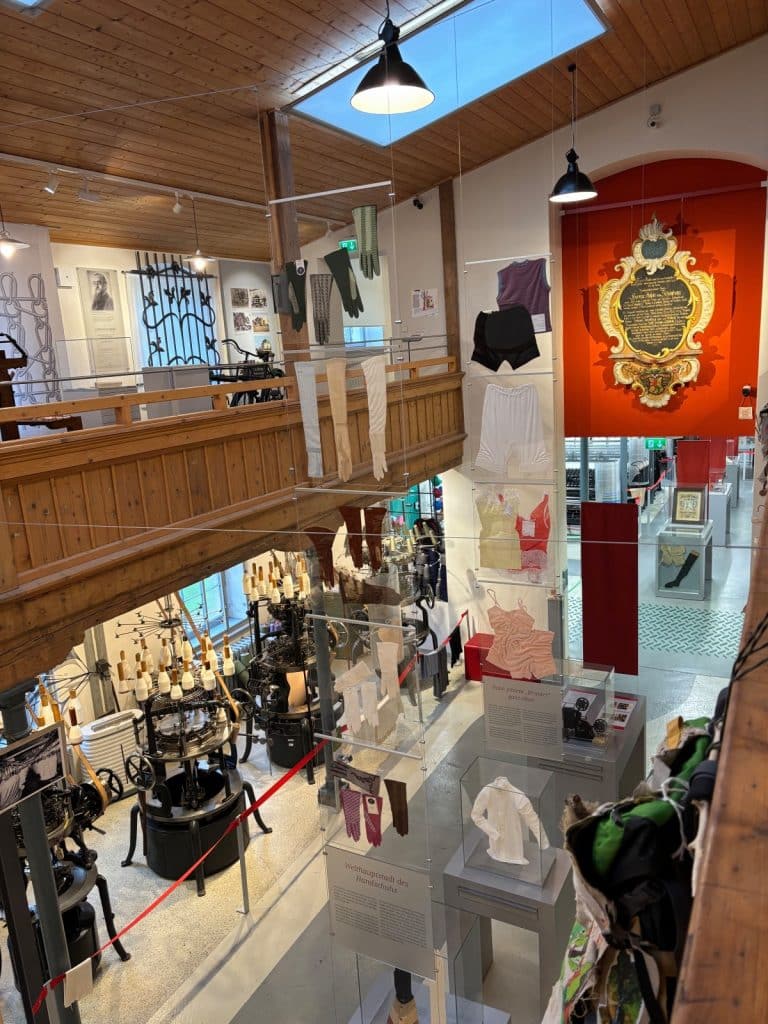 Blick von einer hölzernen Empore in den Ausstellungsraum des Esche-Museums in Sachsen, in dem historische Textilmaschinen sowie an Glaswänden präsentierte Handschuhe und Unterwäsche zu sehen sind.