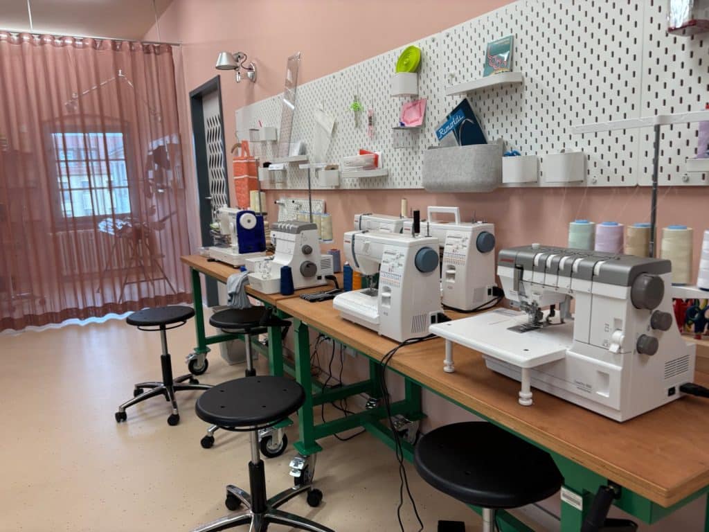 Ein modernes Textillabor im Esche-Museum in Sachsen mit mehreren Nähmaschinen und Overlock-Maschinen auf langen Holztischen vor einer weiß bestückten Lochwand. Schwarze Rollhocker stehen an den Arbeitsplätzen bereit, während im Hintergrund ein rosafarbener Vorhang den Raum abtrennt.