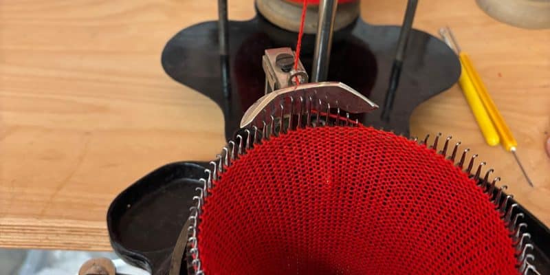 Eine Nahaufnahme zeigt eine historische Rundstrickmaschine im Esche-Museum in Sachsen, die ein rotes, schlauchförmiges Textil aus feinem Garn fertigt. Die roten Maschen hängen an den metallenen Nadeln des runden Strickzylinders, während im Hintergrund eine Garnrolle auf einem Halter zu sehen ist.