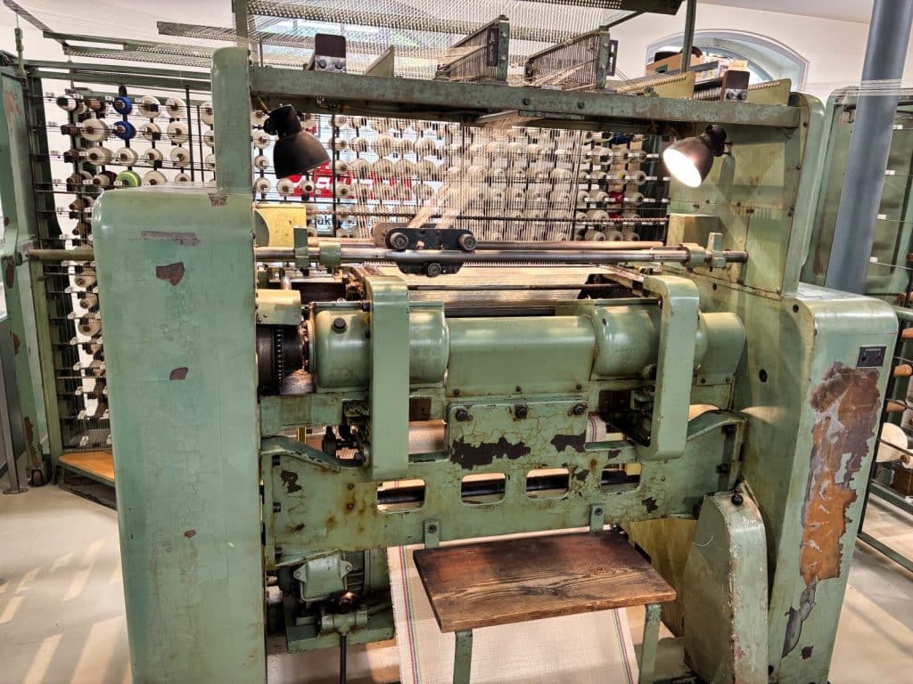 Eine historische, hellgrüne Malimo-Nähwirkmaschine im Esche-Museum in Sachsen, die mit zahlreichen Garnrollen im Hintergrund und einer hölzernen Ablagefläche im Vordergrund ausgestattet ist.