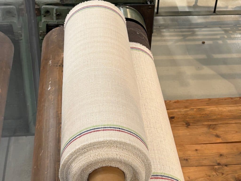 Eine Rolle des in Sachsen entwickelten Malimo-Stoffes mit charakteristischen farbigen Randstreifen liegt auf einer hölzernen Walze im Esche-Museum.
