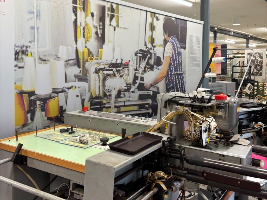 Eine historische Textilmaschine im Esche-Museum in Sachsen steht im Vordergrund vor einer großformatigen Fotowand, die eine Arbeiterin an einer ähnlichen Maschine zeigt.