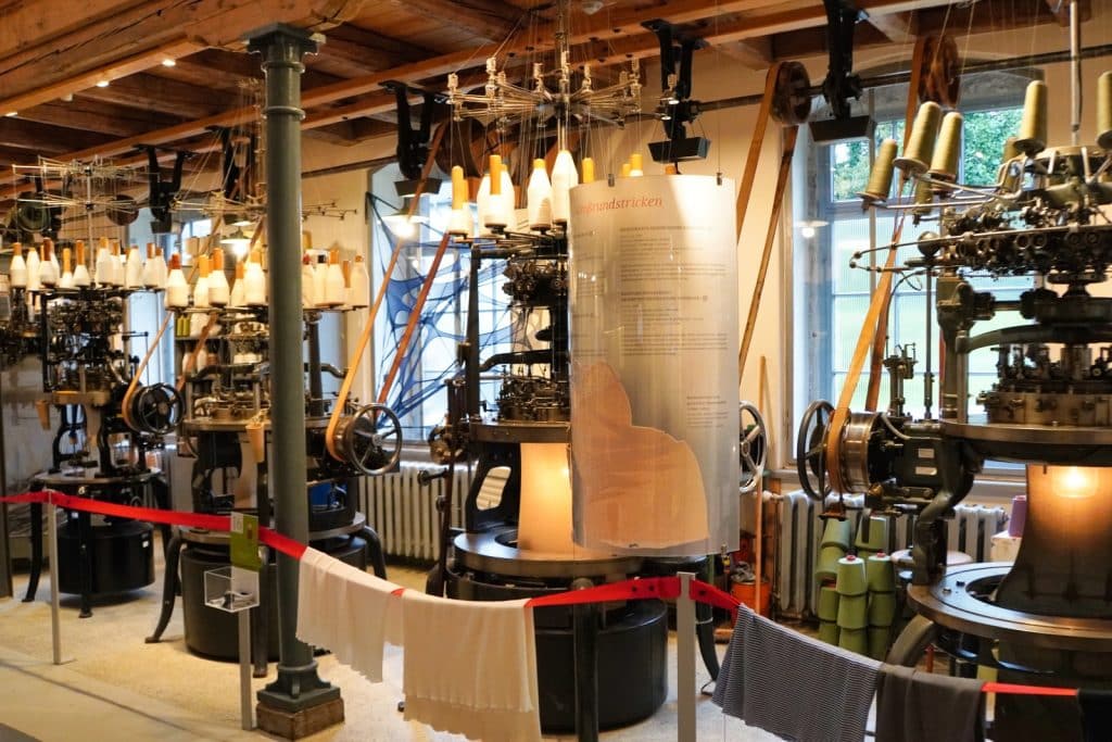 Historische Rundstrickmaschinen im Esche-Museum in Sachsen, die mit zahlreichen Garnspulen bestückt sind und an denen fertige Strickwaren zur Veranschaulichung hängen.