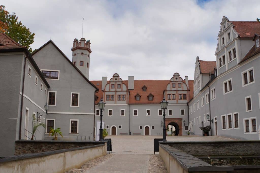 Blick in den gepflasterten Innenhof des Schlosses Forderglauchau in Sachsen mit seinen grauen Fassaden, roten Ziegeldächern und einem markanten weißen Turm unter bewölktem Himmel.