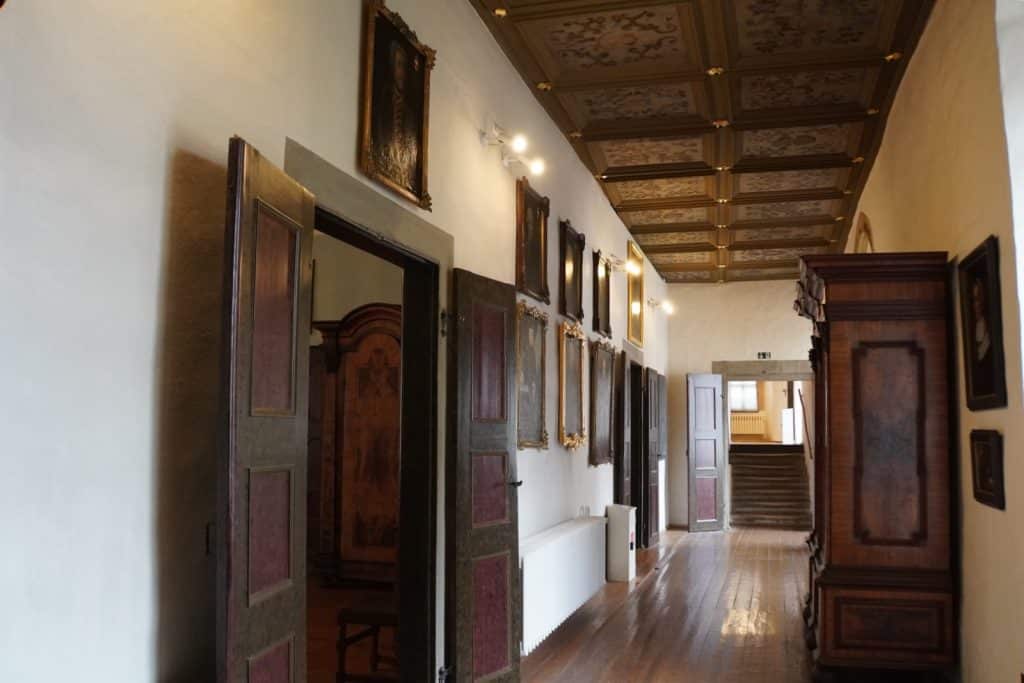 Ein langer, historischer Gang im Schloss Glauchau in Sachsen mit dunklen Holzdielen, einer prachtvollen Kassettendecke und zahlreichen Porträtgemälden an der weißen Wand. Auf der rechten Seite steht ein massiver Holzschrank, während am Ende des Flurs eine Treppe und eine geöffnete Tür zu sehen sind.