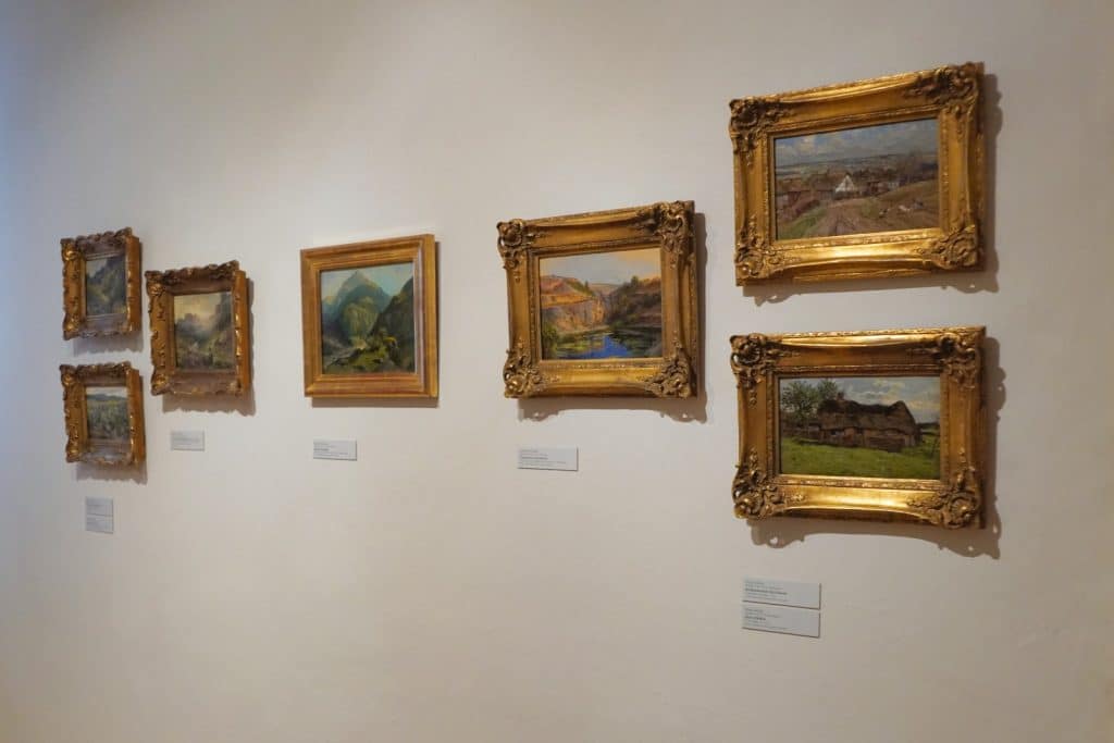 Eine Wanderausstellung in Glauchau, Sachsen, zeigt eine Reihe von gerahmten Landschaftsgemälden in prunkvollen Goldrahmen, die ordentlich nebeneinander an einer weißen Galeriewand hängen.