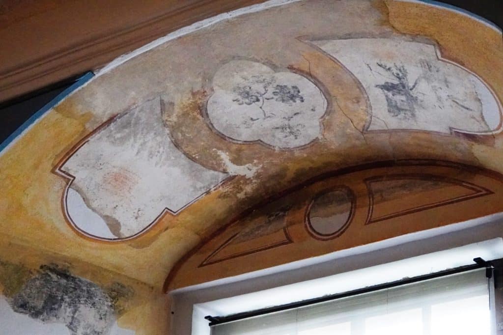 Historische Wandmalereien in einer Fensternische im Schloss Glauchau, Sachsen, zeigen florale und landschaftliche Motive in geschwungenen Kartuschen auf ockerfarbenem Grund. Die Malereien sind teilweise verblasst und weisen deutliche Altersspuren auf dem Putz auf.