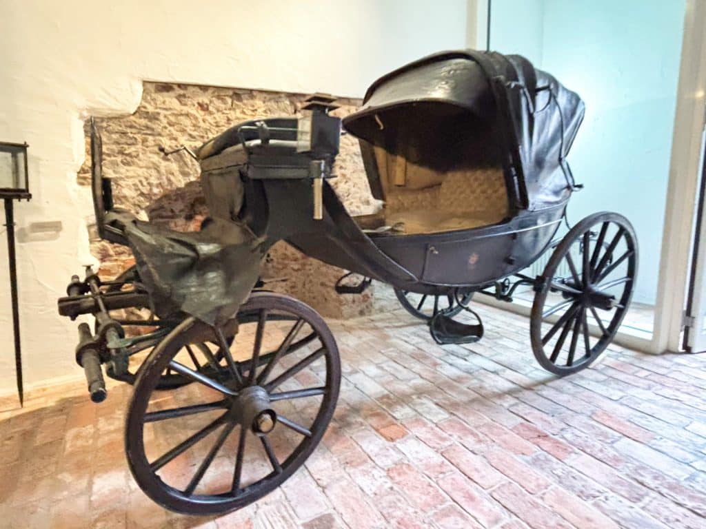 Eine historische schwarze Kutsche aus Sachsen steht in einem Ausstellungsraum in Glauchau auf einem Ziegelboden vor einer rustikalen Steinwand.
