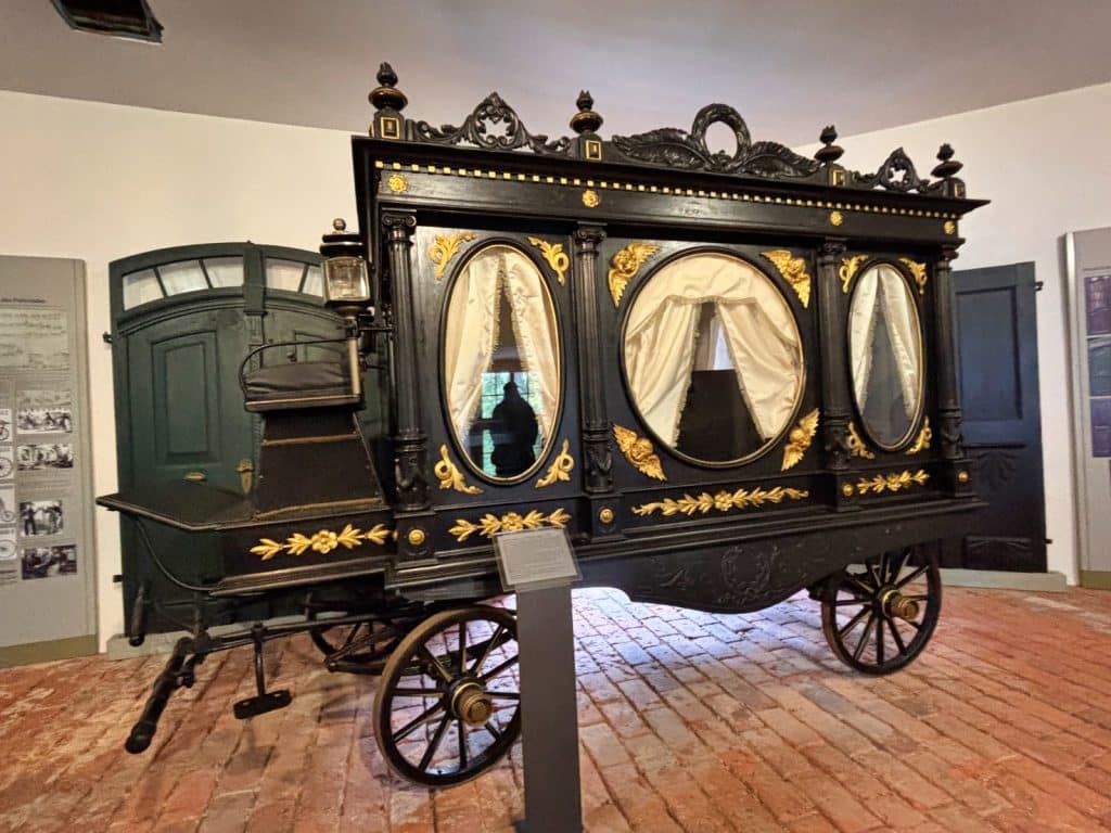 Ein prunkvoller, historischer Leichenwagen aus Glauchau, Sachsen, steht auf einem Ziegelboden in einem Museum und ist mit aufwendigen schwarzen Holzschnitzereien, goldenen Ornamenten und ovalen Fenstern mit weißen Vorhängen verziert.