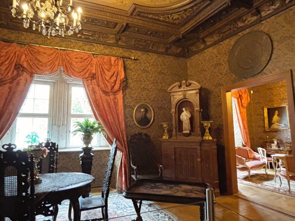 Ein prunkvoller, historischer Raum im Schloss Glauchau in Sachsen mit gemusterten Tapeten, einer verzierten Holzdecke, schweren rosa Vorhängen an einem großen Fenster und antiken Möbeln wie einem runden Tisch und einem hölzernen Schrein. Durch eine offene Tür ist ein angrenzendes, helleres Zimmer mit weiteren Gemälden und Sitzmöbeln sichtbar.