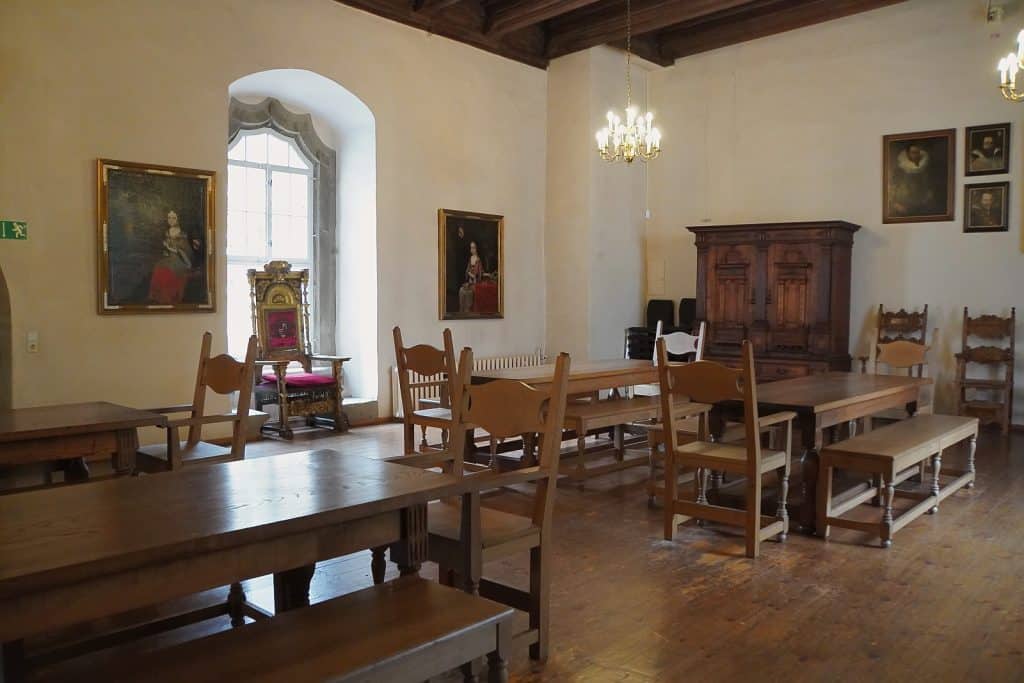Ein historischer Saal im Schloss Glauchau in Sachsen mit dunklen Holzbalkendecken, mehreren massiven Holztischen, Stühlen und einer prunkvollen Thron-Sitzgelegenheit vor einem Fenster. An den hellen Wänden hängen klassische Porträtgemälde, während ein Kronleuchter und ein antiker Holzschrank das rustikale Ambiente ergänzen.