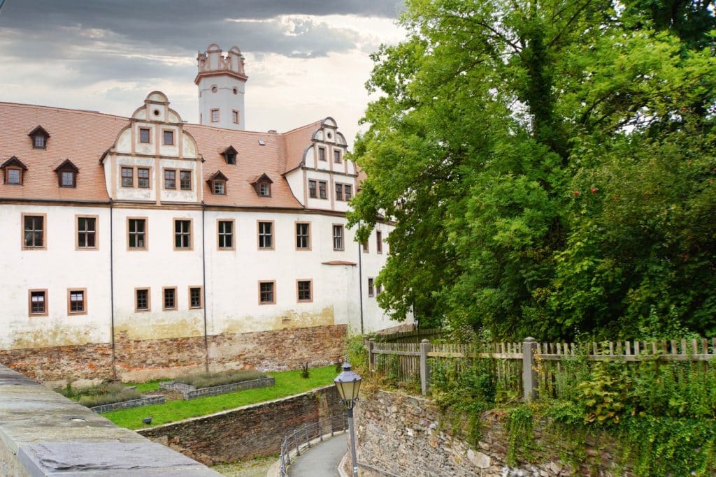 Das Schloss Glauchau in Sachsen mit seiner weißen Fassade, den markanten Renaissance-Giebeln und einem runden Schlossturm hinter einer historischen Steinmauer und dichten grünen Bäumen.