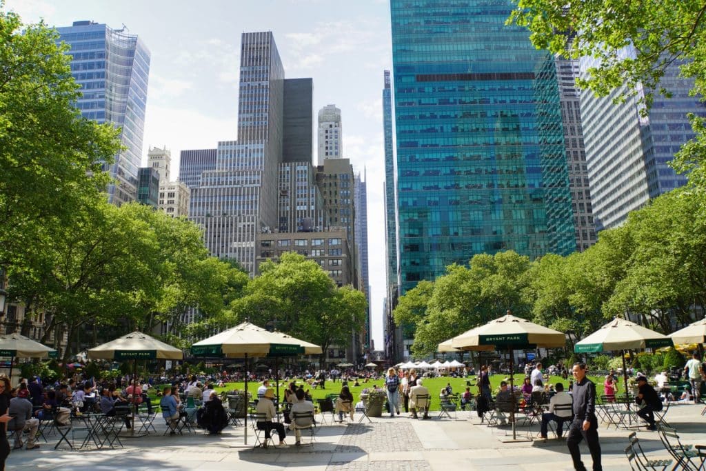 Zahlreiche Menschen entspannen sich an Tischen unter Sonnenschirmen und auf der grünen Liegewiese im Bryant Park in New York, der von dichten Bäumen und hoch aufragenden Wolkenkratzern umgeben ist.