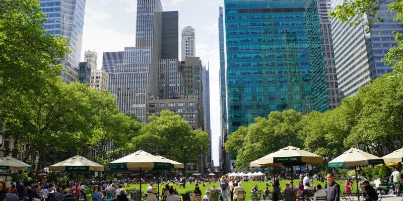 Zahlreiche Menschen entspannen sich an Tischen unter Sonnenschirmen und auf der grünen Liegewiese im Bryant Park in New York, der von dichten Bäumen und hoch aufragenden Wolkenkratzern umgeben ist.