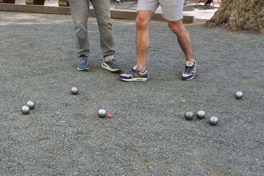 Zwei Personen spielen Boule im New Yorker Bryant Park auf einem Kiesplatz, wobei mehrere silberne Metallkugeln um eine kleine rote Zielkugel verteilt liegen.