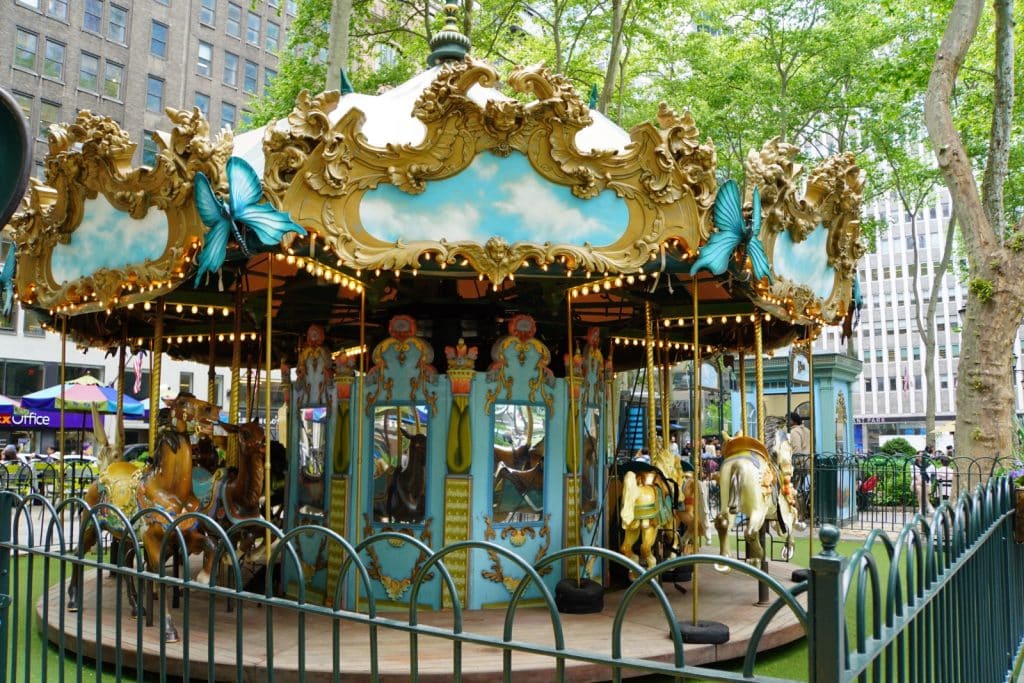 Das festlich dekorierte Le Carrousel im Bryant Park in New York zeigt kunstvoll geschnitzte Tiere und goldene Ornamente unter einem blau bemalten Baldachin mit Schmetterlingsfiguren. Das Karussell ist von einem grünen Metallzaun umgeben und steht vor einer Kulisse aus Stadtgebäuden und grünen Bäumen.