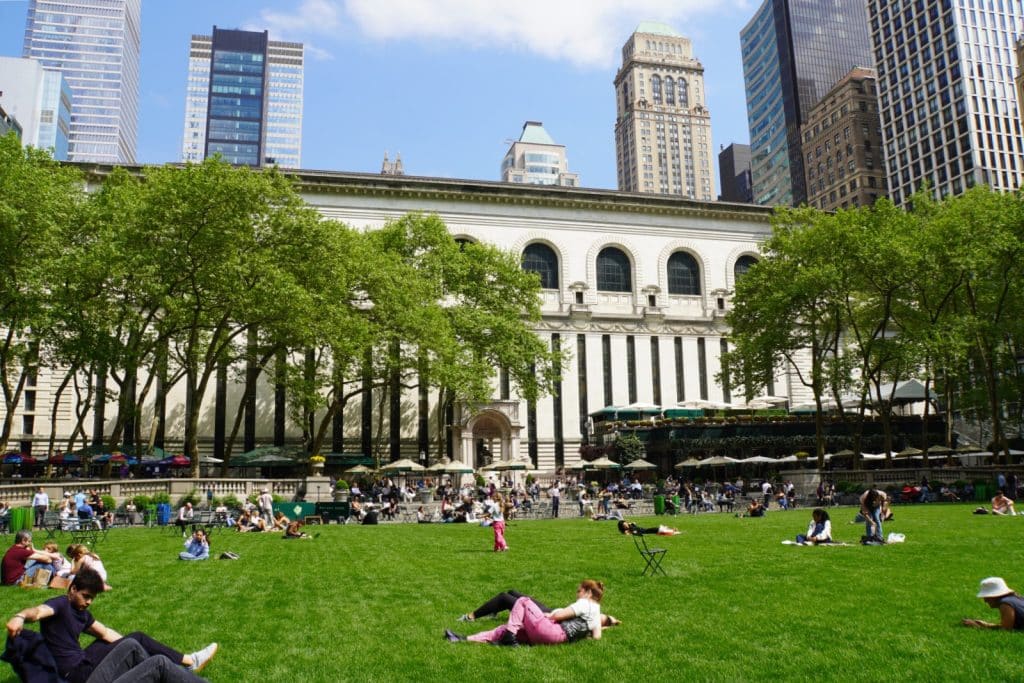 Menschen entspannen auf dem grünen Rasen des Bryant Parks in New York vor der Kulisse der New York Public Library und umliegender Wolkenkratzer.
