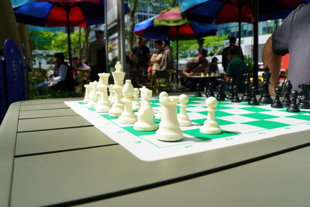 Nahaufnahme eines Schachbretts mit weißen und schwarzen Figuren auf einem Tisch im Bryant Park in New York, während im Hintergrund Menschen unter bunten Sonnenschirmen sitzen.